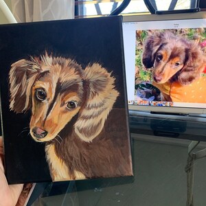 May include: A square canvas painting of a dachshund dog with brown and tan fur, set against a black background. The dog's eyes are brown, and the painting captures the dog's likeness with detailed brushstrokes. The artwork is held by a person.
