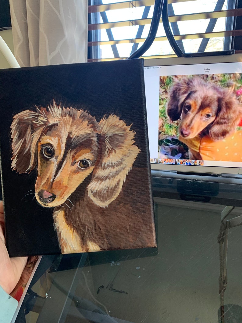 May include: A square canvas painting of a dachshund dog with brown and tan fur, set against a black background. The dog's eyes are brown, and the painting captures the dog's likeness with detailed brushstrokes. The artwork is held by a person.