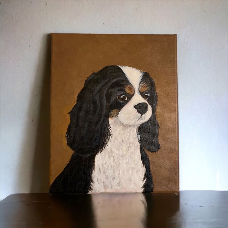 May include: A canvas painting of a Cavalier King Charles Spaniel dog. The dog has black, white, and brown fur, with a focused expression. The background is a solid brown color, and the painting is displayed on a wall.