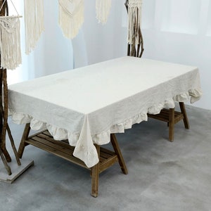 May include: A white linen tablecloth with a ruffled edge, draped over a wooden table with a shelf. The tablecloth is made of a natural fabric and has a rustic look.