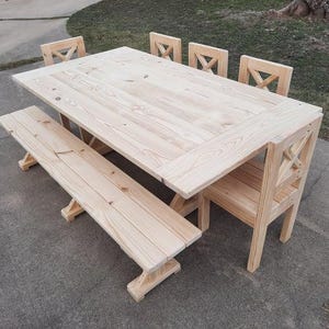 May include: A light-colored wooden dining set consisting of a rectangular table, a long bench, and three chairs. Each chair features a distinct X-shaped backrest design. The furniture is constructed from natural wood with visible grain patterns.