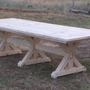 May include: A large, rectangular, light-colored wooden dining table featuring a smooth, flat tabletop surface. The table is supported by three distinct X-shaped trestle bases connected by a central horizontal beam, all crafted from natural wood.