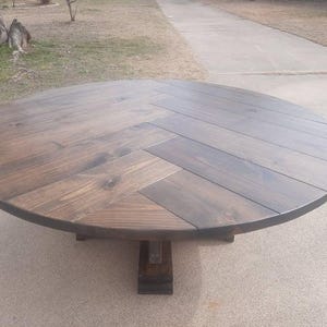 May include: A round wooden table featuring a dark brown stained finish and a prominent herringbone pattern across the tabletop surface. The table is supported by a central pedestal base constructed from matching dark wood planks.