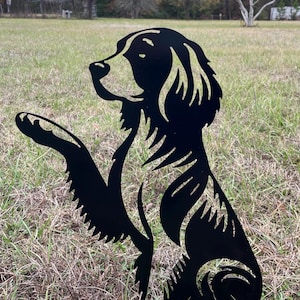 May include: A black metal silhouette of a dog sitting with its paw raised. The dog is depicted in a stylized, artistic manner, with cut-out details. The artwork is placed outdoors on a grassy surface.