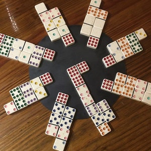 May include: A set of dominoes with colorful dots arranged in a circular pattern on a wooden surface. The dominoes are made of white plastic with black lines separating the dots. The dots are red, green, blue, yellow, and orange.