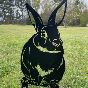 May include: Black metal silhouette of a rabbit sitting on a grassy lawn. The rabbit has large ears and a detailed face.