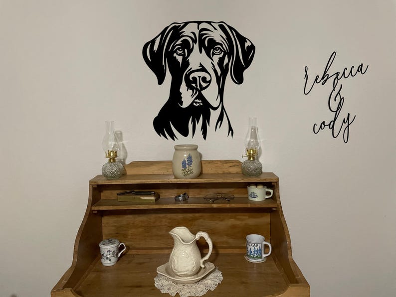 May include: A black and white silhouette of a dog's head with the names "Rebecca & Cody" written in cursive on a white wall. A wooden shelf with two oil lamps, a pitcher, and mugs is in the foreground.