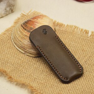 May include: A dark brown leather case with a decorative stitched border. The case has a rounded top and a small embossed design. It rests on a burlap surface, with a seashell and a piece of quartz in the background.