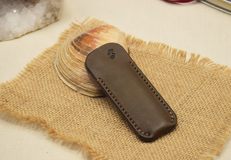 May include: A dark brown leather case with a decorative stitched border. The case has a rounded top and a small embossed design. It rests on a burlap surface, with a seashell and a piece of quartz in the background.