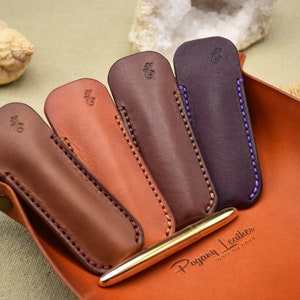 May include: Four leather pen cases in shades of brown, with contrasting stitching and a gold pen. The cases are arranged in a leather tray, with the brand name "Pogany Leather" visible. The cases have a small embossed design.