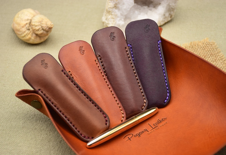 May include: Four leather pen cases in shades of brown, with contrasting stitching and a gold pen. The cases are arranged in a leather tray, with the brand name "Pogany Leather" visible. The cases have a small embossed design.