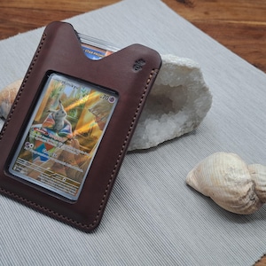 May include: A brown leather card holder with a clear window, holding a colorful trading card. The holder is on a gray surface, next to a seashell and a geode. The card features a cartoon character.