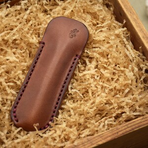 May include: A brown leather case with a decorative stitched edge and a small embossed design. The case is displayed in a wooden box filled with shredded paper. The case is designed to hold a small object.