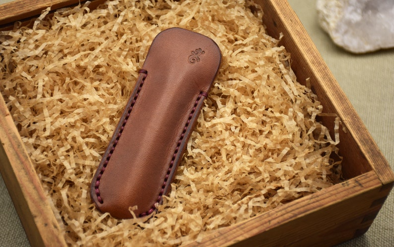 May include: A brown leather case with a decorative stitched edge and a small embossed design. The case is displayed in a wooden box filled with shredded paper. The case is designed to hold a small object.
