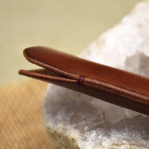 May include: Close-up of a brown leather case, possibly for glasses or a pen. The case is stitched with a contrasting purple thread. The leather has a smooth texture and a warm, inviting color. The case is resting on a white and gray stone.