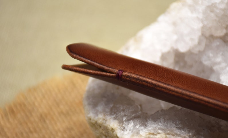 May include: Close-up of a brown leather case, possibly for glasses or a pen. The case is stitched with a contrasting purple thread. The leather has a smooth texture and a warm, inviting color. The case is resting on a white and gray stone.