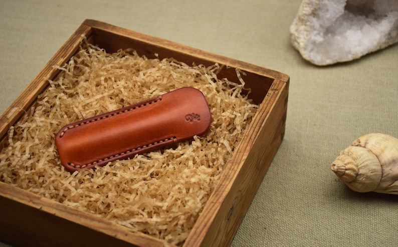 May include: A brown leather knife sheath with stitched edges rests in a wooden box filled with shredded paper. The sheath has a small embossed design. The box sits on a neutral-toned surface.