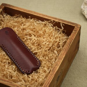 May include: A burgundy leather knife sheath with a stitched border, nestled in a wooden box filled with shredded paper. The sheath has a small embossed design. The box is on a neutral background with a seashell and a rock.