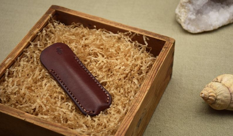 May include: A burgundy leather knife sheath with a stitched border, nestled in a wooden box filled with shredded paper. The sheath has a small embossed design. The box is on a neutral background with a seashell and a rock.
