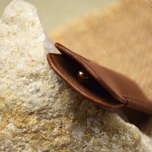 May include: A brown leather case, open to reveal a gold-colored object, possibly a ring or small jewelry item. The case is resting on a rough, light-colored stone, with a textured background.