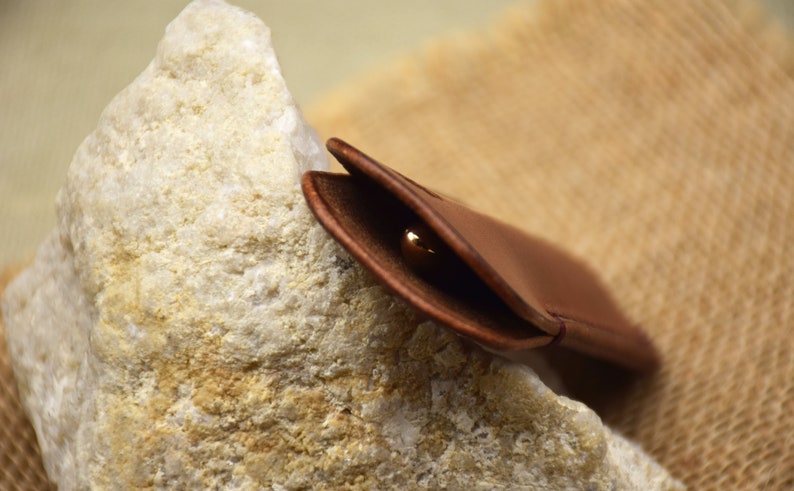 May include: A brown leather case, open to reveal a gold-colored object, possibly a ring or small jewelry item. The case is resting on a rough, light-colored stone, with a textured background.
