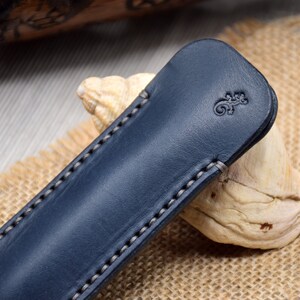 May include: A navy blue leather pen case with a lizard emblem embossed on the front. The case is rectangular with rounded edges and white stitching. It rests on a piece of driftwood and a burlap surface.