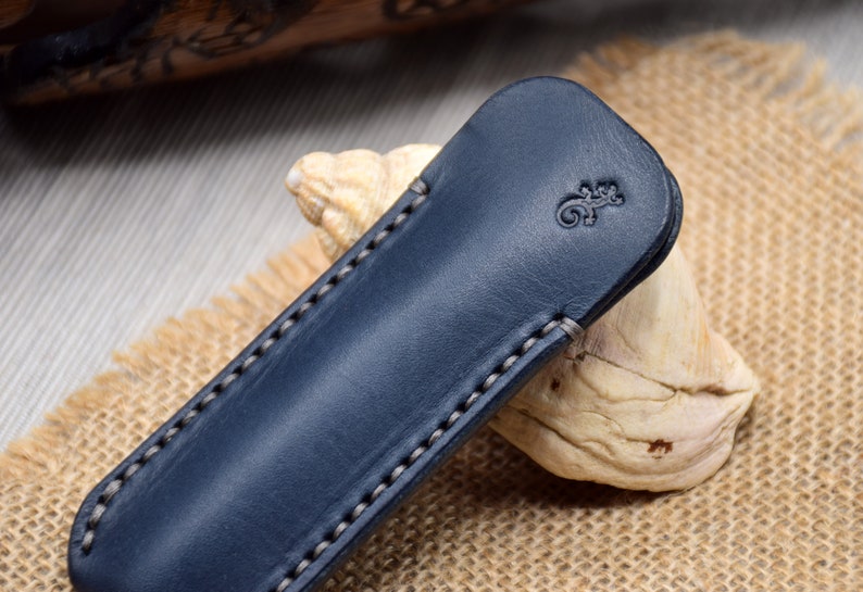 May include: A navy blue leather pen case with a lizard emblem embossed on the front. The case is rectangular with rounded edges and white stitching. It rests on a piece of driftwood and a burlap surface.