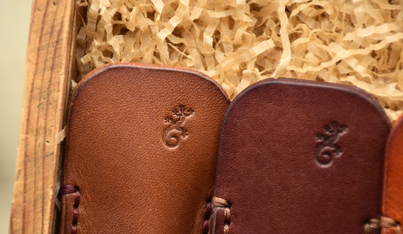 May include: Close-up of several leather items in shades of brown, each with a small embossed lizard design. The items are nestled in a wooden box filled with shredded paper. The leather has a textured appearance.