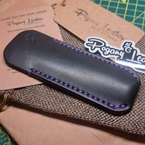 May include: Black leather pen case with purple stitching, resting on a brown fabric pouch. The case is next to a brown tag and a sticker with the text "Pogany Leather". The tag reads "Pogany Leather born by hand".