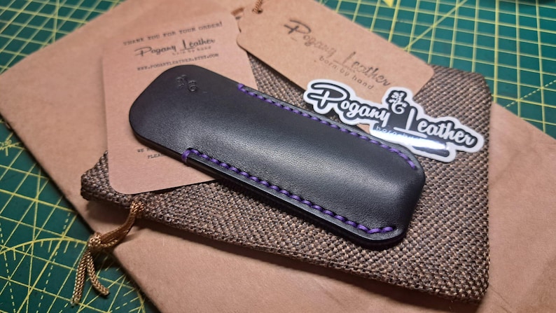 May include: Black leather pen case with purple stitching, resting on a brown fabric pouch. The case is next to a brown tag and a sticker with the text "Pogany Leather". The tag reads "Pogany Leather born by hand".