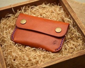 May include: A brown leather wallet with a flap secured by two brass-colored snaps. The wallet has purple stitching and a small embossed design. It rests on shredded paper inside a wooden box.