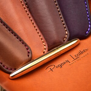 May include: A gold pen rests on an orange leather surface, with the embossed text "Pogany Leather born by hand." Four leather pen sleeves in shades of brown and purple are visible, each with contrasting stitching.