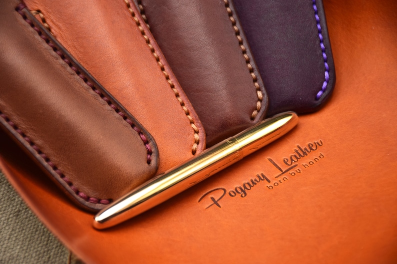 May include: A gold pen rests on an orange leather surface, with the embossed text "Pogany Leather born by hand." Four leather pen sleeves in shades of brown and purple are visible, each with contrasting stitching.