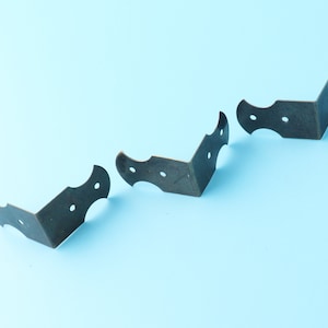Könnte beinhalten: Drei antike, bronzefarbene Eckwinkel mit dekorativen Kanten und jeweils drei kleinen Löchern. Die Winkel sind diagonal auf einem hellblauen Hintergrund angeordnet, geeignet für Möbel oder Bastelprojekte.