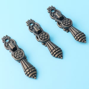 May include: Three antique bronze drawer pulls with ornate floral designs. The pulls have a decorative, slightly raised, circular center with a floral pattern. The pulls are on a light blue background.