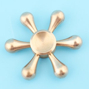 Op de afbeelding: Een gouden metalen fidget spinner met zes armen. De armen zijn traanvormig en het midden van de spinner is rond.