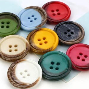 Könnte beinhalten: Sortiment von zehn bunten Plastikknöpfen mit jeweils vier Löchern. Die Knöpfe sind in verschiedenen Farben, darunter Grün, Blau, Rot, Gelb, Braun und Schwarz. Die Knöpfe sind rund und haben eine glatte, glänzende Oberfläche.