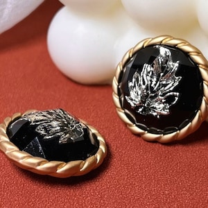 以下が含まれることがあります： 銀の葉のデザインが施された黒と金のボタン2つ。ボタンはロープのような縁取りがされている。