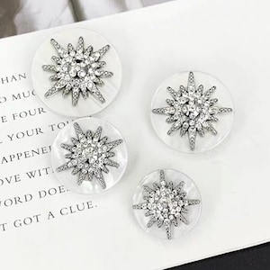 Puede incluir: Cuatro botones redondos blancos con un diseño de estrella plateada. Cada estrella está hecha de diamantes de imitación.