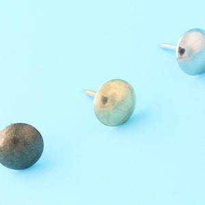 Könnte beinhalten: Drei runde Nägel in verschiedenen Ausführungen: Silber, Gold und Antik-Bronze. Die Nägel sind in einer Reihe auf einem hellblauen Hintergrund angeordnet.