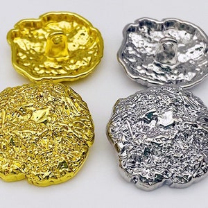 Pode incluir: Dois conjuntos de botões decorativos, um conjunto é dourado e o outro é prateado. Os botões têm uma superfície texturizada irregular e um centro elevado.