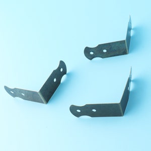 Puede incluir: Tres soportes de esquina de latón antiguo con bordes decorativos y dos agujeros a cada lado. Los soportes están sobre un fondo azul claro.