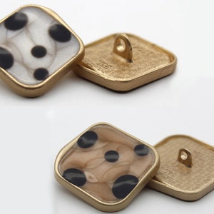 Op de afbeelding: Twee vierkante knopen met een gouden metalen frame en een zwart-witte stippenpatroon. De knopen hebben een lus aan de achterkant om te naaien.