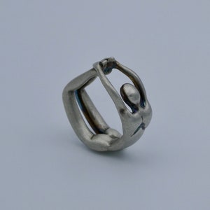 May include: A silver ring featuring a stylized human figure in a yoga pose. The figure is sculpted in a three-dimensional design, with the arms raised above the head and the body bent backwards.