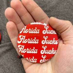 May include: Red round button with the text "Florida Sucks" repeated in white cursive lettering.
