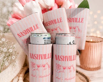 May include: A set of light pink and white striped foam can coolers featuring the text Nashville, illustrations of cowboy boots, martini glasses, and guitars, and the script text Madi's Bachelorette. Two silver aluminum cans are inserted inside.