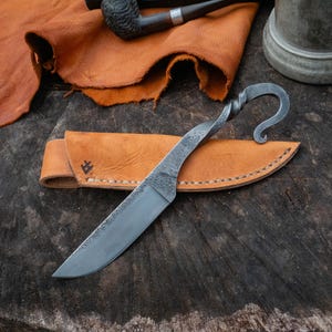 Pu&ograve; includere: Un coltello forgiato a mano con un manico attorcigliato e un'estremit&agrave; curva, su un fodero in pelle marrone chiaro. La lama &egrave; argentata e il fodero &egrave; cucito con un motivo decorativo. Il coltello &egrave; esposto su una superficie di legno rustico.