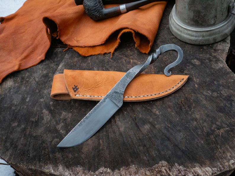 Pu&ograve; includere: Un coltello forgiato a mano con un manico attorcigliato e un'estremit&agrave; curva, su un fodero in pelle marrone chiaro. La lama &egrave; argentata e il fodero &egrave; cucito con un motivo decorativo. Il coltello &egrave; esposto su una superficie di legno rustico.