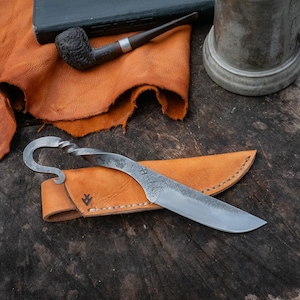 Pu&ograve; includere: Un coltello forgiato a mano con un manico curvo e un fodero in pelle. La lama &egrave; argentata e il fodero &egrave; marrone chiaro. Sono visibili anche una pipa, un libro e una tazza di peltro.
