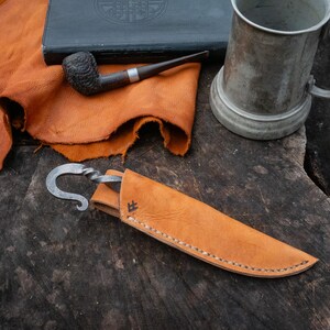 Pu&ograve; includere: Un coltello artigianale con una lama curva e un fodero in pelle. Il coltello &egrave; in metallo e ha un manico attorcigliato. Il fodero &egrave; marrone chiaro e cucito. Sono raffigurati anche una pipa, un boccale di peltro e una pelle di cuoio.